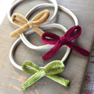 Op de afbeelding: Drie fluwelen haarbandjes met strikken in verschillende kleuren: mosterdgeel, bordeauxrood en olijfgroen. De haarbandjes zijn gemaakt van een zacht, rekbaar materiaal en perfect om elke outfit een vleugje kleur te geven.