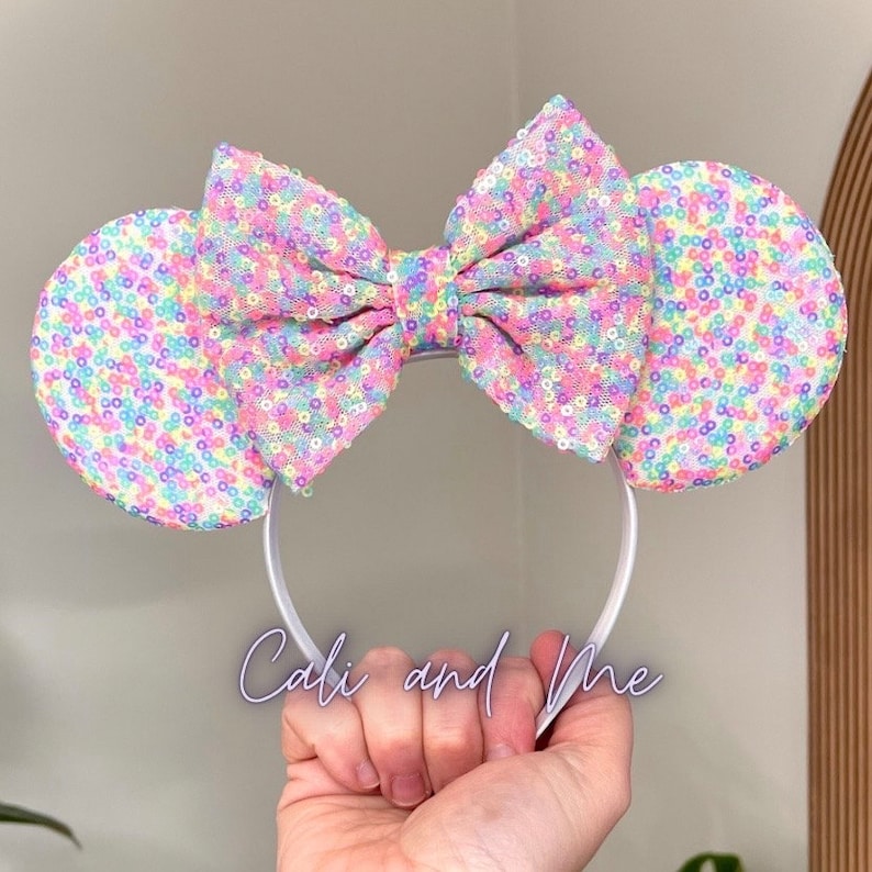 K&ouml;nnte beinhalten: Pastellfarbene Regenbogen-Sequin-Minnie-Maus-Ohren-Haarreif mit einer gro&szlig;en Schleife. Der Haarreif ist wei&szlig;.