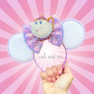 May include: A whimsical headband featuring a teapot character with a bow made of sequins in shades of pink and purple. The ears are white with a beaded trim. The headband is held up against a pink background with the text "cali and me".