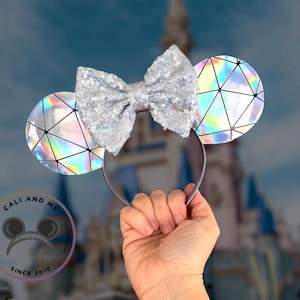 May include: Silver sequined bow and ears headband. The ears have a geometric design with iridescent accents. The headband is held up against a blurred background of a castle and sky.