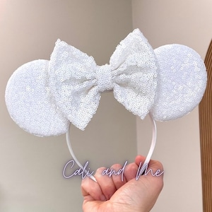 May include: White sequined Minnie Mouse ears headband with a large white bow. The headband is held in a hand with the text "Cali and Me" visible below.