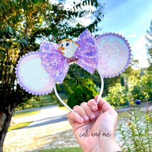 May include: A white headband with sequined mouse ears and a large purple sequined bow. The bow features a character figurine. The ears are white with a lavender trim. The headband is held up against a background of trees and a blue sky.