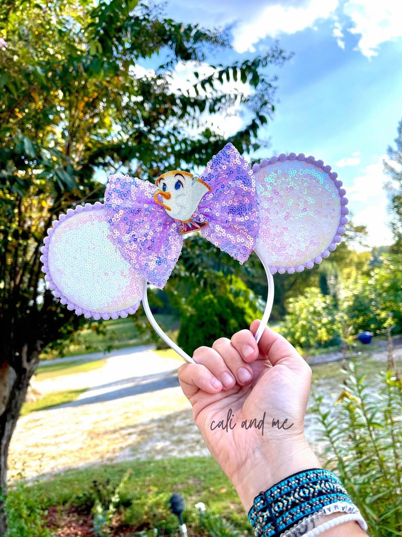 May include: A white headband with sequined mouse ears and a large purple sequined bow. The bow features a character figurine. The ears are white with a lavender trim. The headband is held up against a background of trees and a blue sky.