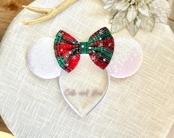 May include: A festive headband featuring a red and green plaid bow with white snowflake accents. The headband includes white sequined ears and a clear plastic band. The text "Callie and Me" is visible on the band.