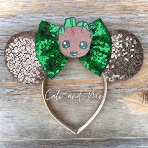 May include: Green and brown sequined Minnie Mouse ears with a brown and green Groot character in the center. The headband is gold.