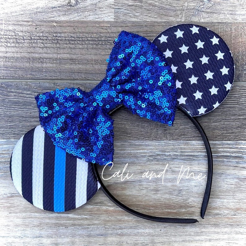 May include: Blue and white mouse ears with a blue sequined bow. The ears feature a white and blue striped pattern and a blue background with white stars.