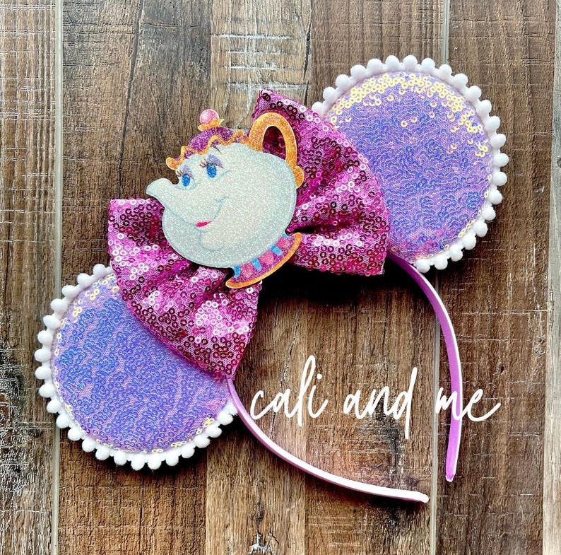 May include: A purple headband with sequined ears and a large bow. The bow features a cartoon teapot character. The ears are purple with white trim. The bow is a mix of pink and purple sequins. The text "cali and me" is written on the headband.