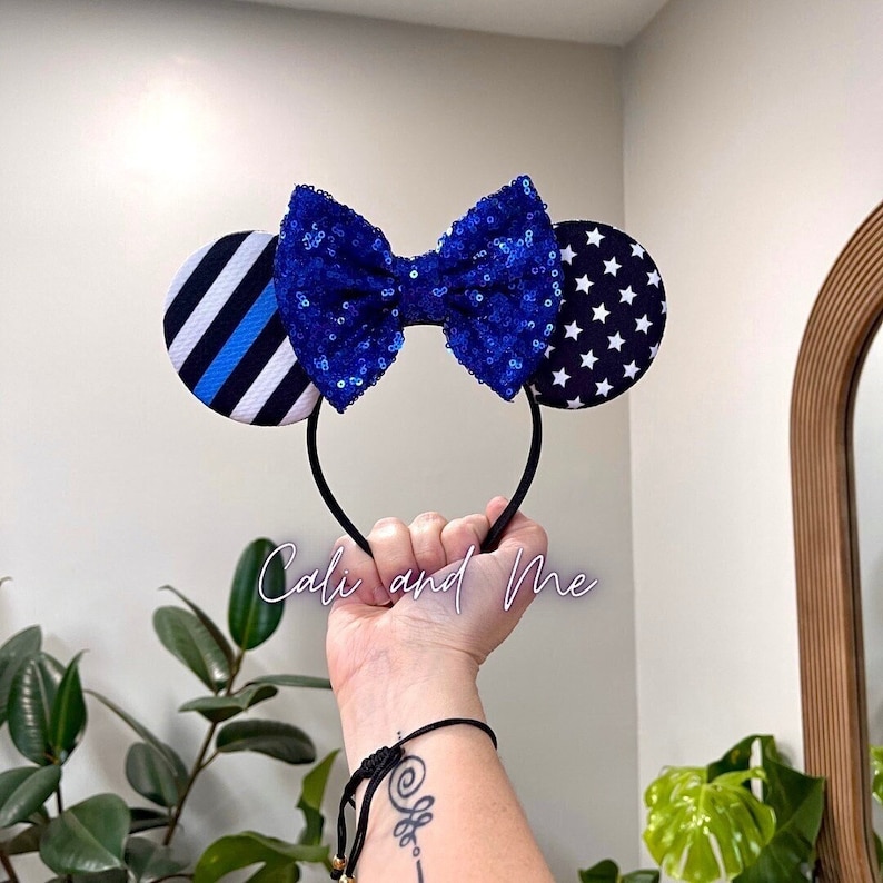 May include: Blue and white mouse ears with a blue sequin bow. The left ear has a black and white striped pattern with a blue line. The right ear has a white background with black stars. The headband is black.