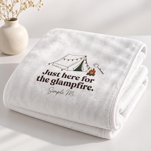 May include: A white fleece blanket featuring a graphic of a camping tent with string lights, a campfire, and a marshmallow on a stick. The text reads: Just here for the glampfire. Simple Me. The blanket is folded neatly on a flat surface.