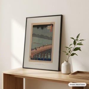 Hiroshige's Sudden Shower framed print in a black frame, leaning on a wooden sideboard beside a plant