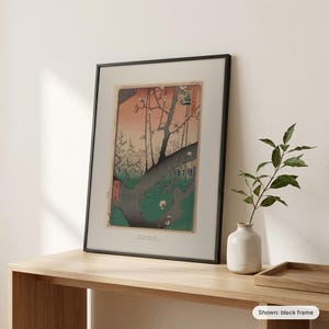Hiroshige's Plum Park in Kameido framed print in a black frame, leaning on a wooden sideboard beside a plant