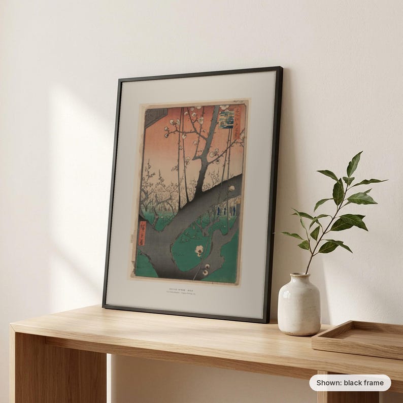 Hiroshige's Plum Park in Kameido framed print in a black frame, leaning on a wooden sideboard beside a plant