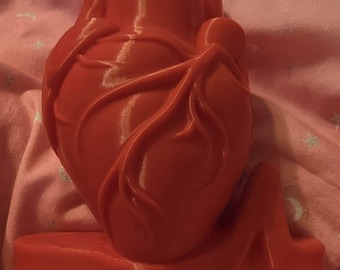 Può includere: Una scultura di cuore anatomico rosso brillante in tre dimensioni, caratterizzata da una superficie liscia modellata con dettagli di vene in rilievo. Il cuore è fissato su una base orizzontale piatta che si estende verso destra.
