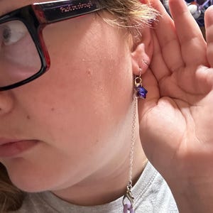 Pu&ograve; includere: Un orecchino color argento con un piccolo ciondolo a forma di stella blu scuro sfaccettato, fissato a una lunga e delicata catenina argentata. La catena termina con un moschettone che sostiene due piccoli anelli o perline circolari lilla.
