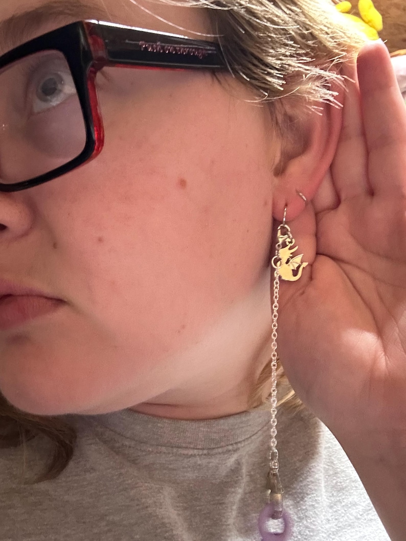 Pu&ograve; includere: Un orecchino color argento con un piccolo ciondolo a forma di drago piatto color oro, fissato a una delicata catenella metallica. La catena scende verso il basso e si collega a un anello circolare viola chiaro, creando un lungo accessorio pendente.