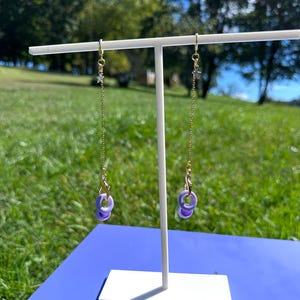Pu&ograve; includere: Un paio di orecchini lunghi pendenti caratterizzati da una sottile catenina color oro fissata a un piccolo ciondolo a forma di stella color argento. La parte inferiore di ogni catena &egrave; collegata a un moschettone dorato che sostiene due piccoli anelli circolari color viola chiaro.