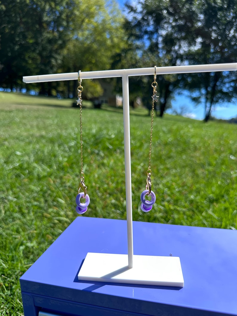 Pu&ograve; includere: Un paio di orecchini lunghi pendenti caratterizzati da una sottile catenina color oro fissata a un piccolo ciondolo a forma di stella color argento. La parte inferiore di ogni catena &egrave; collegata a un moschettone dorato che sostiene due piccoli anelli circolari color viola chiaro.