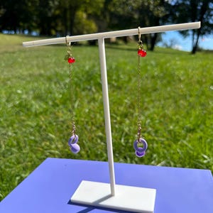 Pu&ograve; includere: Un paio di orecchini pendenti lunghi con catenina dorata, un piccolo ciondolo a forma di ciliegia rossa con foglia verde e un ciondolo a forma di kettlebell viola chiaro alla base. Gli orecchini sono su un supporto a T bianco all'aperto.