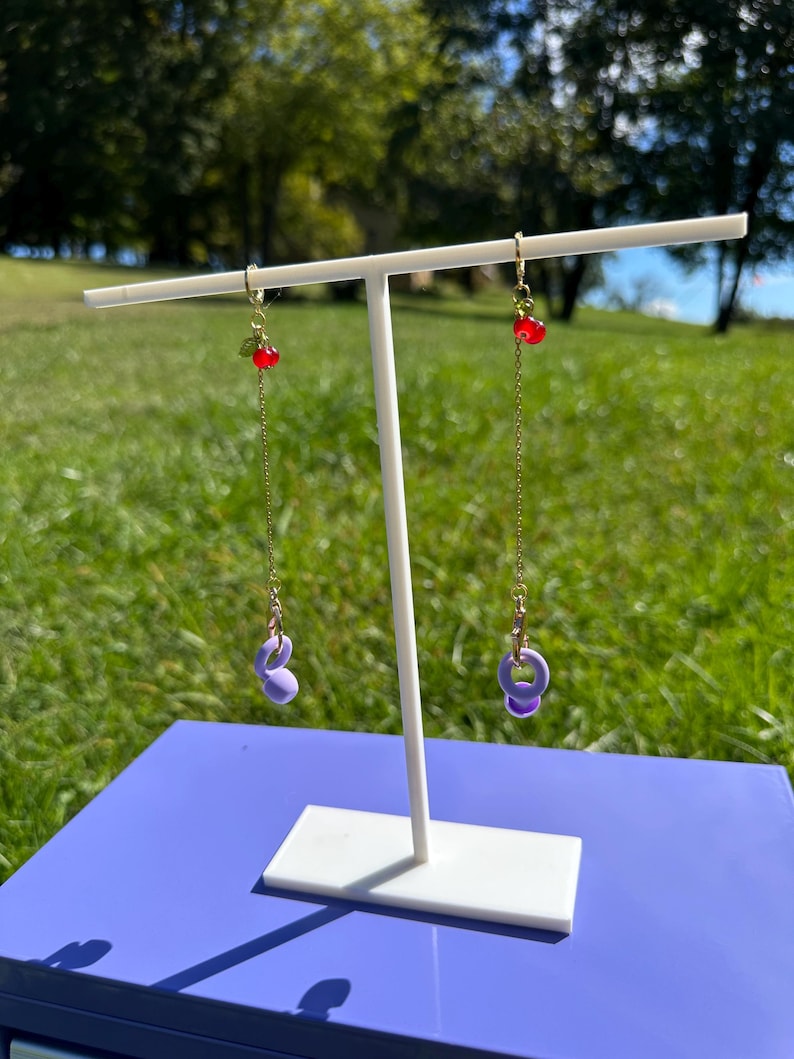 Pu&ograve; includere: Un paio di orecchini pendenti lunghi con catenina dorata, un piccolo ciondolo a forma di ciliegia rossa con foglia verde e un ciondolo a forma di kettlebell viola chiaro alla base. Gli orecchini sono su un supporto a T bianco all'aperto.