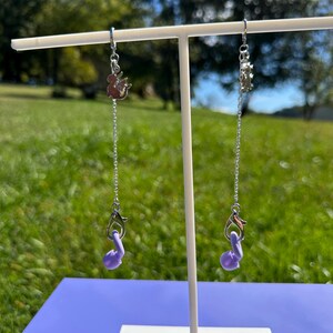 Pu&ograve; includere: Una coppia di catenelle in metallo color argento appese a un espositore bianco. Ogni catenella presenta un piccolo ciondolo a forma di drago in argento in alto e un supporto per auricolari in silicone lilla fissato a un moschettone argentato.