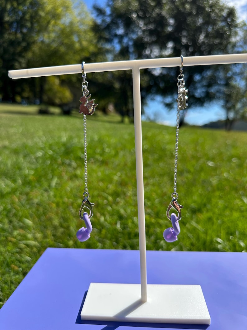 Pu&ograve; includere: Una coppia di catenelle in metallo color argento appese a un espositore bianco. Ogni catenella presenta un piccolo ciondolo a forma di drago in argento in alto e un supporto per auricolari in silicone lilla fissato a un moschettone argentato.
