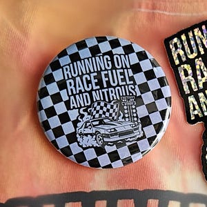 May include: A round button with a black and white checkered pattern and the text "RUNNING ON RACE FUEL AND NITROUS". The button features a graphic of a car and a checkered flag.