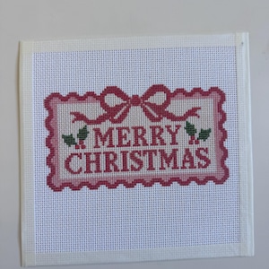 May include: A square white needlepoint canvas featuring a centered rectangular design with a scalloped pink border. The design includes a pink bow at the top, green holly leaves with red berries, and the red text MERRY CHRISTMAS.