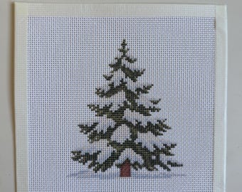 May include: A square piece of white needlepoint canvas featuring a cross-stitch design of a dark green pine tree with a small brown trunk, set against a light blue base representing snow, with white tape binding the edges of the fabric.