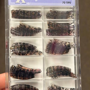 May include: A clear plastic storage case containing 70 pre-designed nail tips. The tips are dark, translucent, and feature a black mottled pattern with gold glitter accents. The label at the top reads Scent Xperience Pre-Designed Nail Tips 70 TIPS.