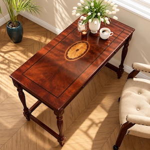 May include: A rectangular wooden desk with a dark mahogany finish, featuring a central oval inlay with a sunburst pattern and black border lines. The desk stands on four turned legs and is accompanied by a beige upholstered armchair.