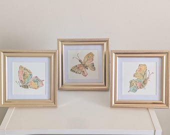 May include: Three square wall art pieces featuring watercolor butterfly illustrations. Each artwork is encased in a metallic gold frame with a white mat border. The butterflies display soft hues of pink, yellow, blue, and brown on paper.