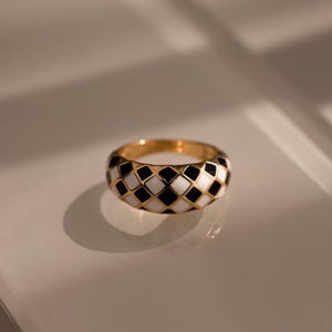 May include: A gold-toned ring featuring a repeating diamond-shaped checkerboard pattern in alternating black and white enamel. The band has a rounded, domed profile with a polished finish, showcasing a consistent grid of small, square-like shapes.
