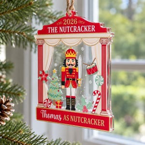 May include: A clear acrylic Christmas ornament shaped like a theater stage featuring a nutcracker, a Christmas tree, a drum, and a candy cane. The top text reads 2026 THE NUTCRACKER and the bottom text reads Thomas AS NUTCRACKER.