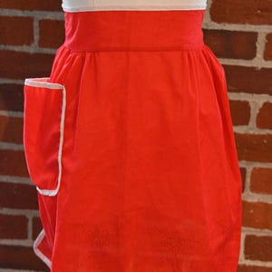 May include: A bright red apron featuring a wide waistband, a single side pocket, and crisp white trim along the edges. The apron is gathered at the waist and displays a subtle, tonal embroidered pattern on the lower section of the fabric.
