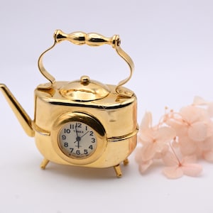 May include: A gold-toned metal clock shaped like a miniature teapot with a handle, spout, and four small legs. The clock face is circular with black numerals and the word Timex printed in the center. The clock is set on a plain surface.