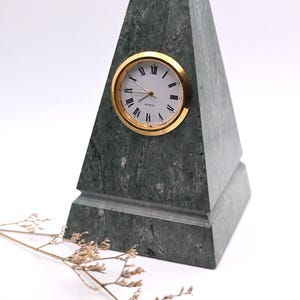 May include: A pyramid-shaped desk clock crafted from dark gray mottled stone. The clock face is circular with a gold-toned rim, white background, black Roman numerals, and the word Quartz printed in black. The clock is set into the stone.