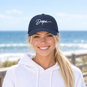 May include: A navy blue baseball cap featuring the word Dope embroidered in white cursive script across the front panel. The cap has a structured crown, a curved brim, and a button on top, providing a classic look with clean white lettering.