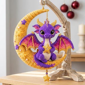 May include: A purple crocheted dragon with large black eyes, pink and yellow wings, and a cream-colored belly, sitting on a yellow crocheted crescent moon. The moon is decorated with small purple stars and white beads, with a gold star hanging below.