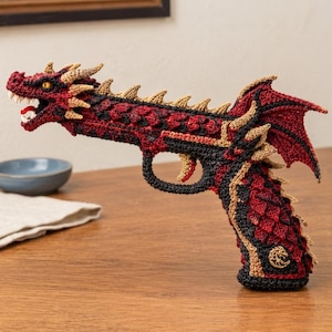 May include: A crocheted sculpture shaped like a handgun with a dragon head at the barrel. The piece features deep red, black, and tan yarn, with a textured scale pattern, a wide-open mouth with white teeth, and a small dragon wing detail.