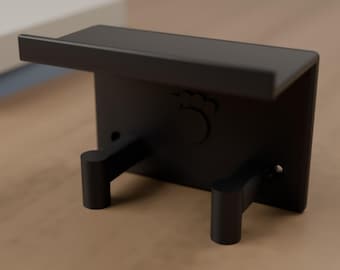 May include: A matte black wall-mounted shelf featuring a small top ledge and two downward-facing cylindrical hooks. The central back panel displays a subtle embossed paw print design, and two small circular screw holes are visible on the sides.