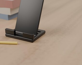 May include: A black rectangular phone stand with a solid upright back panel and two front support tabs. The stand is constructed from a matte black material and features a simple, functional design for holding a mobile device upright.