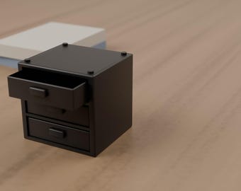 May include: A small, matte black desktop organizer featuring three stacked drawers. The top drawer is pulled open, revealing an empty interior. Each drawer is equipped with a simple, rectangular handle. The unit has a square, boxy shape.