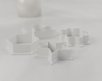 May include: A set of four white plastic cookie cutters in various geometric and organic shapes, including a hexagon, a heart, a star, and a scalloped flower shape, arranged together on a flat surface.