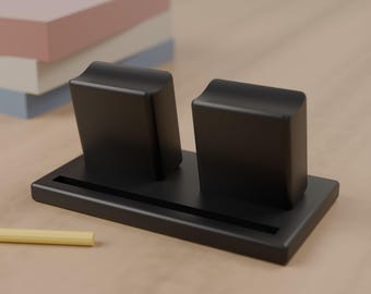 May include: A matte black desk organizer featuring a rectangular base with a long, narrow slot and two upright, rectangular blocks with concave tops. The organizer is positioned on a smooth surface near a yellow pencil and stacked paper.