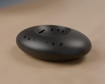 May include: A smooth, matte black, oval-shaped object with a concave center. The top surface features a series of small, thin, rectangular ventilation slits arranged in a circular pattern around the central depression.