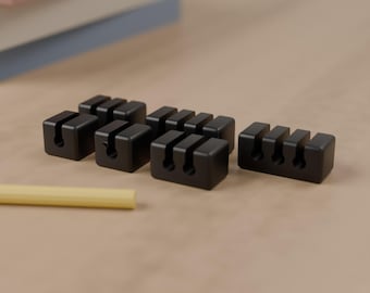 May include: A collection of small black rectangular cable organizers made of a matte material. Each piece features one or more vertical slots designed to hold cords in place. A single yellow cylindrical rod lies nearby on the surface.