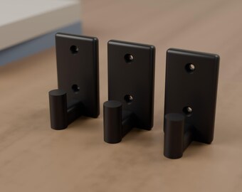 May include: Three matte black wall hooks, each featuring a rectangular mounting plate with two circular screw holes and a cylindrical hook extending outward. These hardware pieces are uniform in design, color, and finish, standing upright.