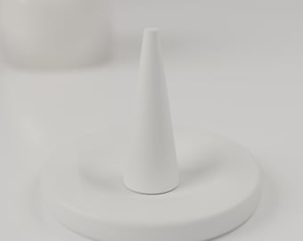 May include: A matte white ring holder featuring a circular base and a central vertical cone. The object is constructed from a solid, smooth material with a uniform finish, designed to hold rings securely on the upright tapered post.