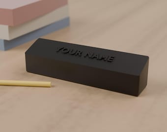 May include: A matte black rectangular desk nameplate featuring the embossed text YOUR NAME on the front face. The object is positioned horizontally with a thin yellow pencil lying parallel to its base on a light wooden surface.