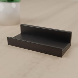 May include: A matte black rectangular display stand featuring a low front ledge and a taller back panel, designed with clean lines and a smooth finish to hold small items securely in an upright position for clear visibility.
